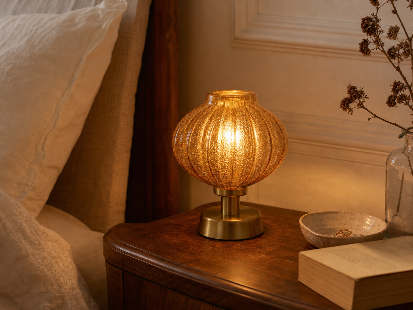 An amber glass vintage-style table lamp casting a warm glow in a softly layered bedroom scene.